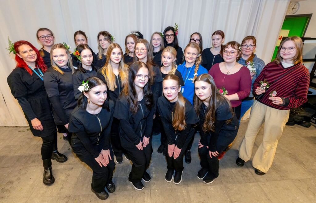 Ryhmä nuoria ja aikuisia seisoo yhdessä sisätiloissa. Suurin osa henkilöistä on pukeutunut mustiin vaatteisiin ja heillä on kukkia hiuksissaan. He hymyilevät kameralle ja seisovat ryhmässä vaalean taustan edessä. Oikealla puolella pari henkilöä pitää käsissään pieniä kukkia.