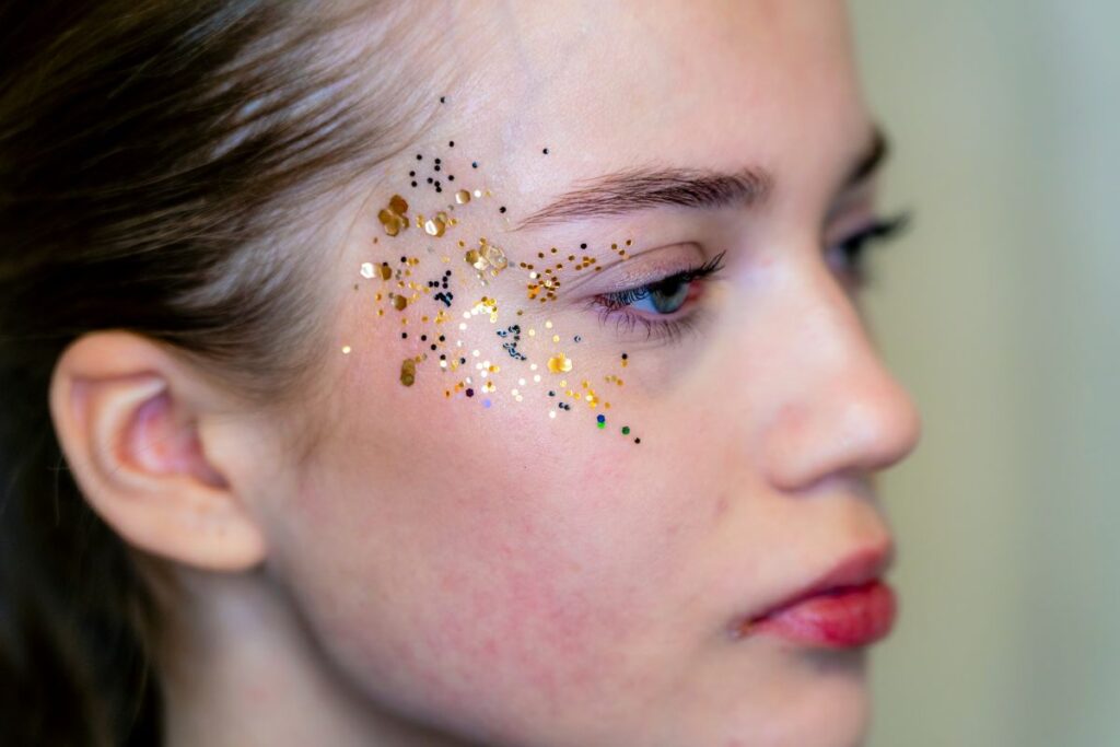 Lähikuva henkilöstä, jolla on kullanvärisiä ja mustia glitterikoristeita oikealla poskellaan. Tausta on epätarkka. Henkilö katsoo oikealle.