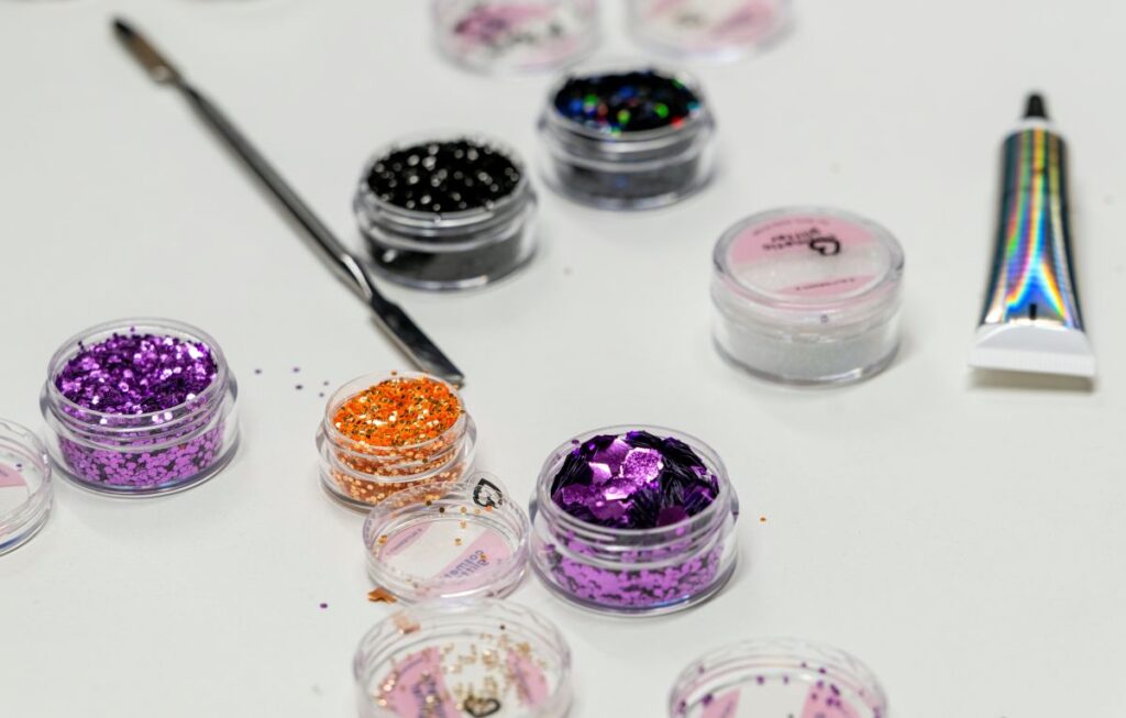 Erilaisia pieniä purkkeja, joissa on värikkäitä glittereitä ja koristeita kynsitaidetta varten. Kuvassa näkyy myös metallinen työkalu ja kiiltävä putkilo valkoisella taustalla.