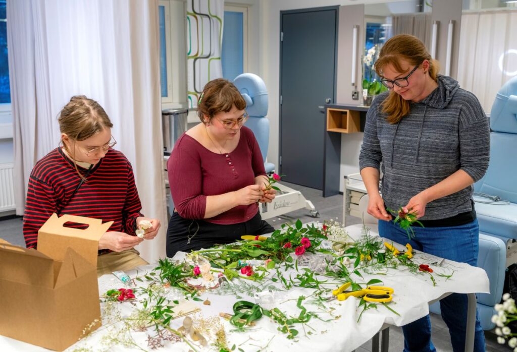 Kolme henkilöä tekee kukka-asetelmia pöydän ääressä sisätiloissa. Pöydällä on erilaisia kukkia ja vihreitä kasveja.