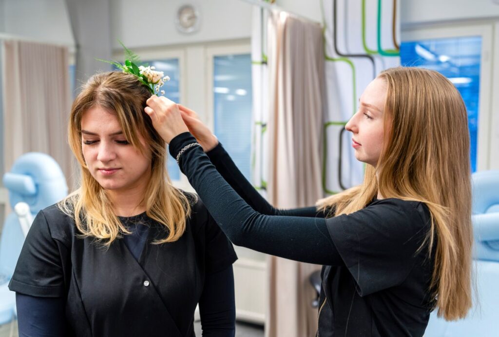 Kaksi naista ammatillisessa ympäristössä, toinen asettaa kukkakoristeen toisen hiuksiin.