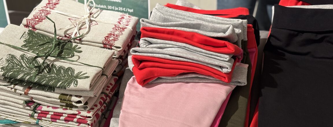 Pöydällä on pino kuvioituja laudeliinoja ja tonttulakkeja. Taustalla näkyy kyltti, jossa lukee "Tekstiili- ja muotialan opiskelijoiden valmistamat tuotteet".