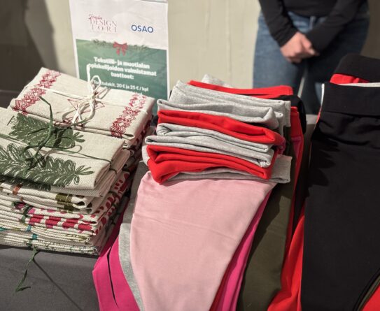 Pöydällä on pino kuvioituja laudeliinoja ja tonttulakkeja. Taustalla näkyy kyltti, jossa lukee "Tekstiili- ja muotialan opiskelijoiden valmistamat tuotteet".