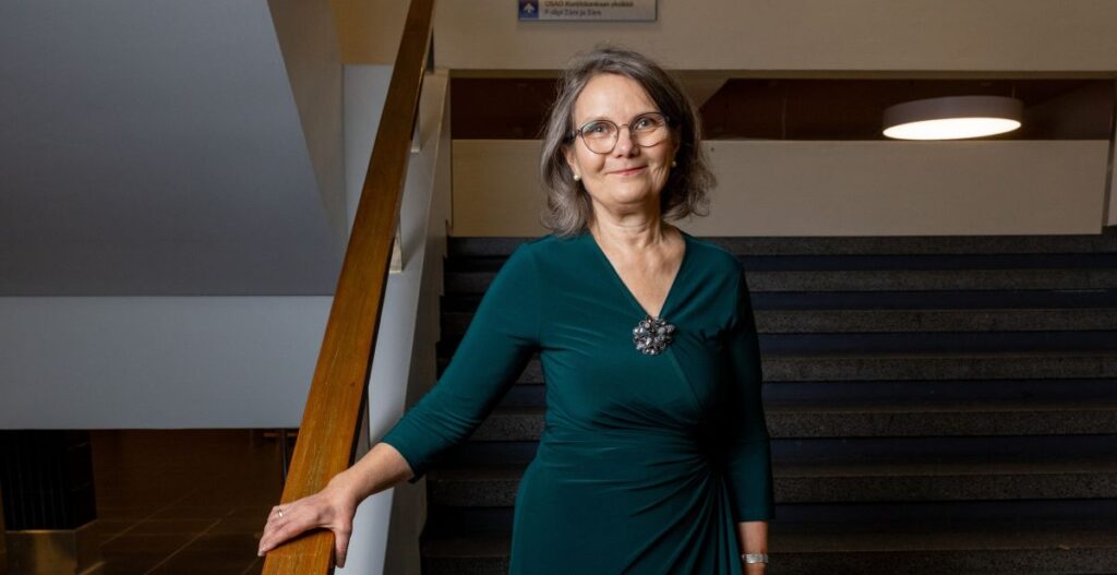 Nainen seisoo portaiden vieressä. Hänellä on yllään vihreä mekko, ja hän hymyilee kameralle. Taustalla on sisätilojen valaistusta ja portaat.