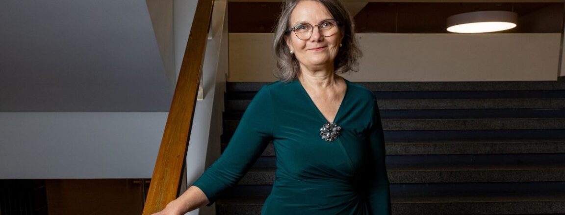 Nainen seisoo portaiden vieressä. Hänellä on yllään vihreä mekko, ja hän hymyilee kameralle. Taustalla on sisätilojen valaistusta ja portaat.