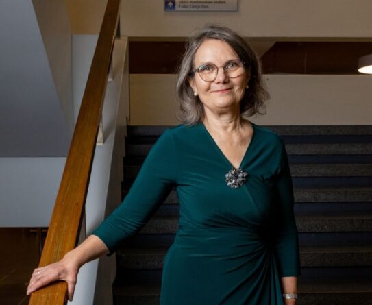 Nainen seisoo portaiden vieressä. Hänellä on yllään vihreä mekko, ja hän hymyilee kameralle. Taustalla on sisätilojen valaistusta ja portaat.