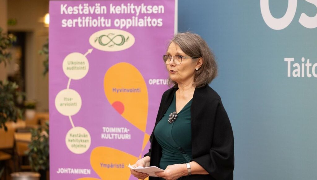 Nainen seisoo puhujana sisätilassa, taustallaan violetti juliste, jossa lukee 'Kestävän kehityksen sertifioitu oppilaitos'. Julisteessa on myös kaavio, jossa mainitaan 'Ulkoinen auditointi', 'Itsearviointi', 'Kestävän kehityksen ohjelma', 'Johtaminen', 'Hyvinvointi', 'Toimintakulttuuri', 'Ympäristö' ja 'Opetus'. Nainen pitää papereita käsissään.