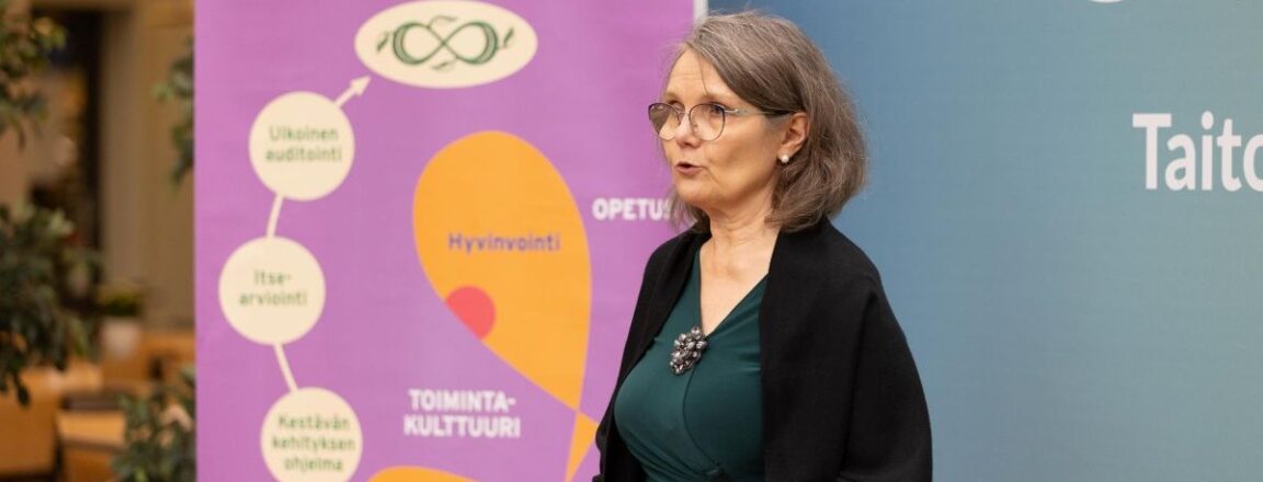 Nainen seisoo puhujana sisätilassa, taustallaan violetti juliste, jossa lukee 'Kestävän kehityksen sertifioitu oppilaitos'. Julisteessa on myös kaavio, jossa mainitaan 'Ulkoinen auditointi', 'Itsearviointi', 'Kestävän kehityksen ohjelma', 'Johtaminen', 'Hyvinvointi', 'Toimintakulttuuri', 'Ympäristö' ja 'Opetus'. Nainen pitää papereita käsissään.