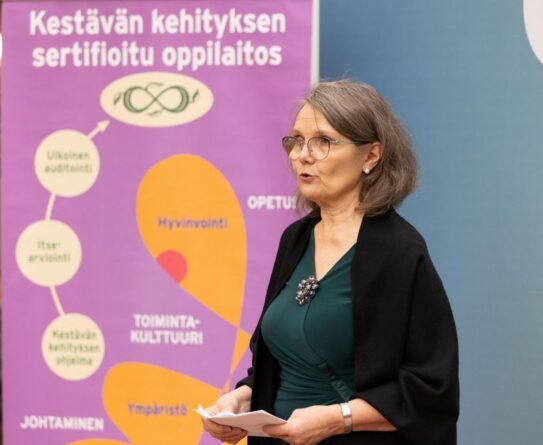 Nainen seisoo puhujana sisätilassa, taustallaan violetti juliste, jossa lukee 'Kestävän kehityksen sertifioitu oppilaitos'. Julisteessa on myös kaavio, jossa mainitaan 'Ulkoinen auditointi', 'Itsearviointi', 'Kestävän kehityksen ohjelma', 'Johtaminen', 'Hyvinvointi', 'Toimintakulttuuri', 'Ympäristö' ja 'Opetus'. Nainen pitää papereita käsissään.