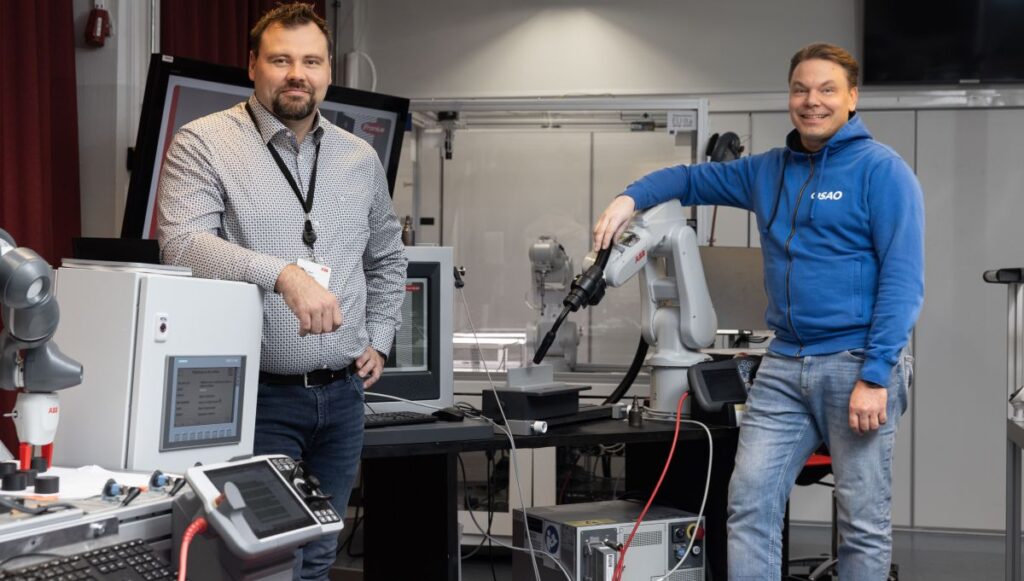 Kaksi henkilöä seisoo laboratoriossa robottien ja tietokoneiden ympäröimänä. Oikealla oleva henkilö nojaa robottikäteen, ja vasemmalla oleva henkilö seisoo kädet ristissä. Molemmat hymyilevät kameralle.