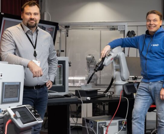 Kaksi henkilöä seisoo laboratoriossa robottien ja tietokoneiden ympäröimänä. Oikealla oleva henkilö nojaa robottikäteen, ja vasemmalla oleva henkilö seisoo kädet ristissä. Molemmat hymyilevät kameralle.