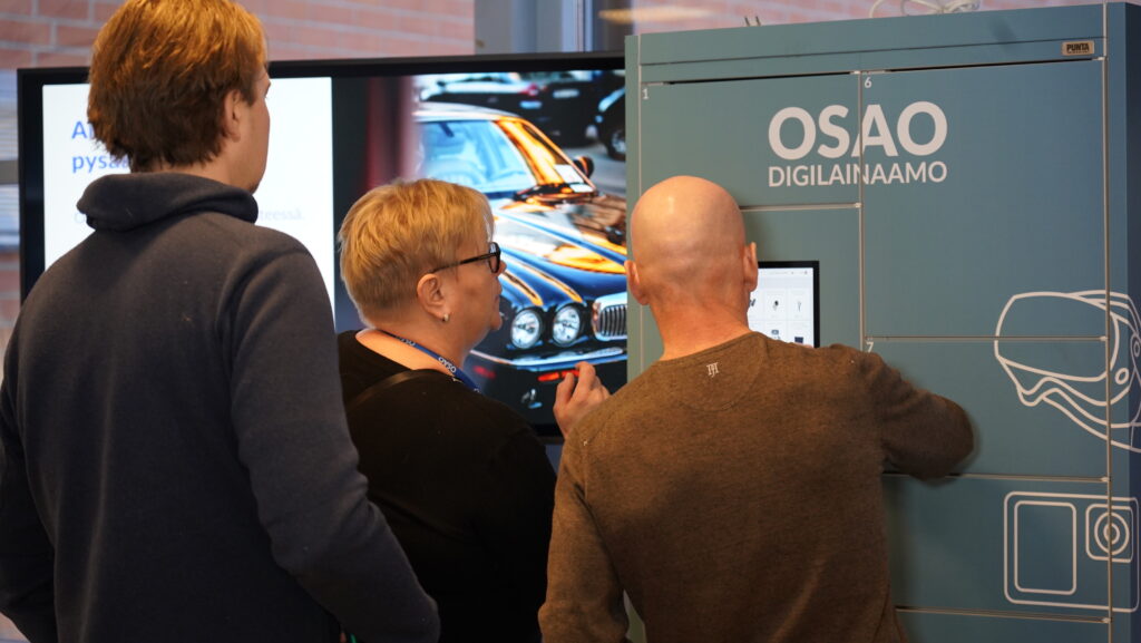 Kolme henkilöä käyttämässä OSAO Digilainaamo-automaattia. Taustalla näkyy näyttö, jossa on kuva autosta.