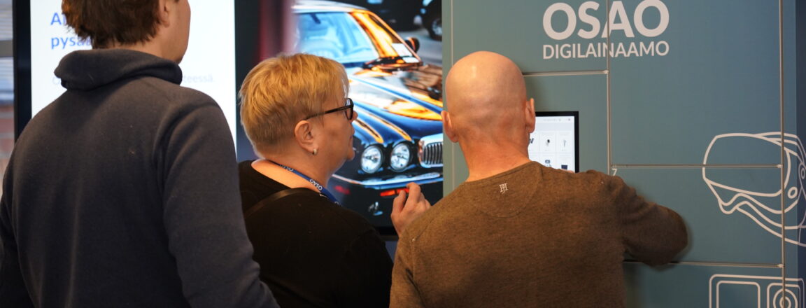 Kolme henkilöä käyttämässä OSAO Digilainaamo-automaattia. Taustalla näkyy näyttö, jossa on kuva autosta.