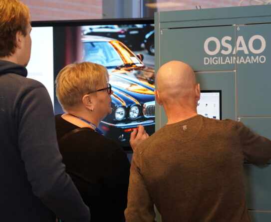 Kolme henkilöä käyttämässä OSAO Digilainaamo-automaattia. Taustalla näkyy näyttö, jossa on kuva autosta.