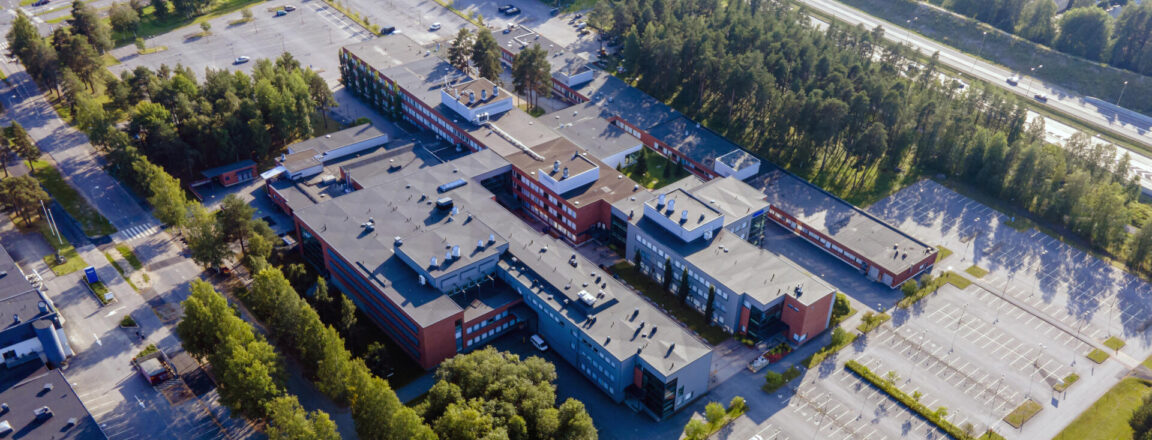 Ilmakuva suuresta koulurakennuksesta, jota ympäröi parkkipaikat ja vehreä metsä. Rakennuksen katolla näkyy useita eri osia ja sen ympäristössä on asfalttiteitä ja pysäköintialueita.