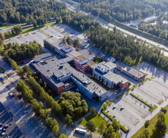 Ilmakuva suuresta koulurakennuksesta, jota ympäröi parkkipaikat ja vehreä metsä. Rakennuksen katolla näkyy useita eri osia ja sen ympäristössä on asfalttiteitä ja pysäköintialueita.