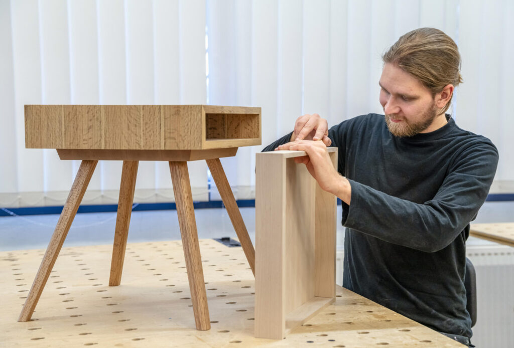 Puuseppä työstämässä puista huonekalua työpajassa. Hänen edessään on pöydällä puinen pöytä, jossa on neljä kartiomaista jalkaa.