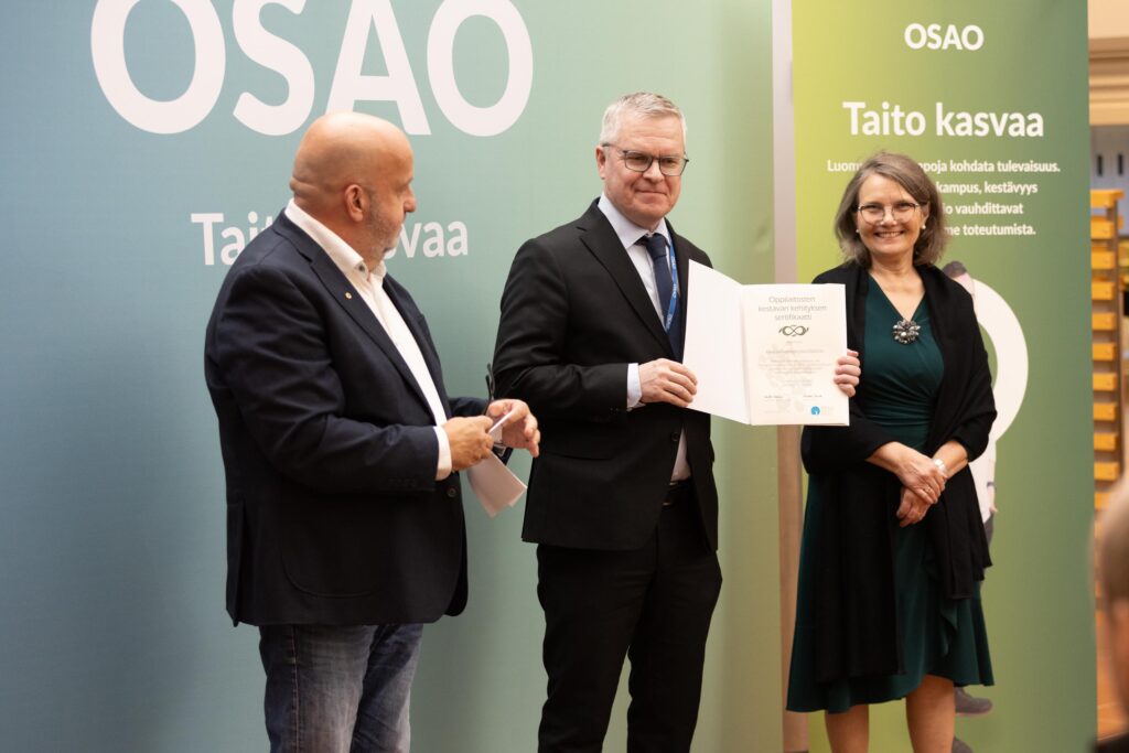 Kolme henkilöä seisoo OSAO-logon edessä. Keskimmäinen henkilö pitelee todistusta. Kaksi muuta henkilöä seisoo hänen vieressään, yksi vasemmalla ja yksi oikealla.