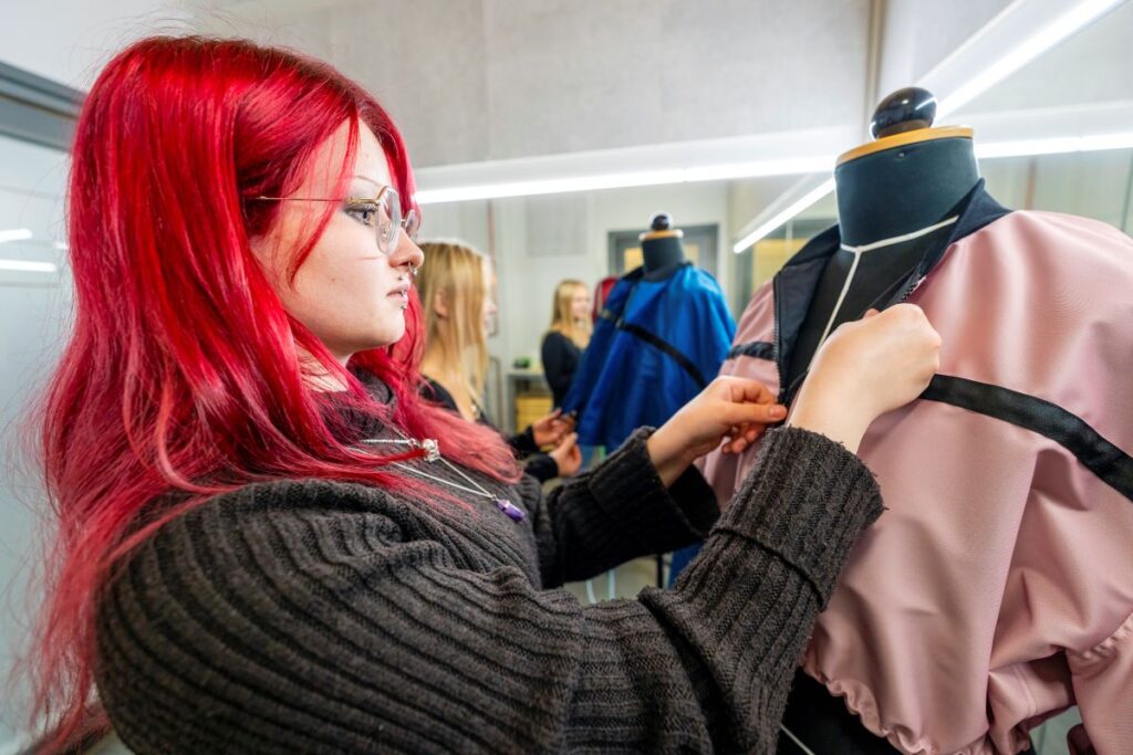 Henkilö, jolla on punaiset hiukset, työskentelee vaatteen parissa muotinäytösnukkeella. Taustalla näkyy toinen henkilö ja sininen vaate. Tilassa on kirkas valaistus.