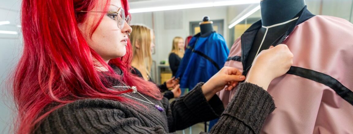 Henkilö, jolla on punaiset hiukset, työskentelee vaatteen parissa muotinäytösnukkeella. Taustalla näkyy toinen henkilö ja sininen vaate. Tilassa on kirkas valaistus.