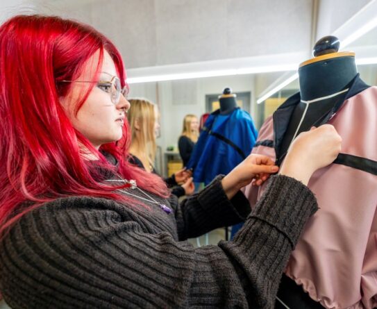 Henkilö, jolla on punaiset hiukset, työskentelee vaatteen parissa muotinäytösnukkeella. Taustalla näkyy toinen henkilö ja sininen vaate. Tilassa on kirkas valaistus.