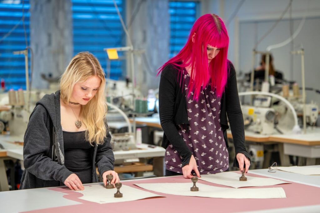 Kaksi henkilöä seisoo ompelimossa ja leikkaa kankaita kaavojen avulla. Taustalla on ompelukoneita.