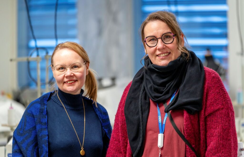 Kaksi hymyilevää naista seisoo yhdessä sisätiloissa. Heillä on silmälasit ja neuleet yllään. Taustalla näkyy ompelukoneita ja ikkunoita.