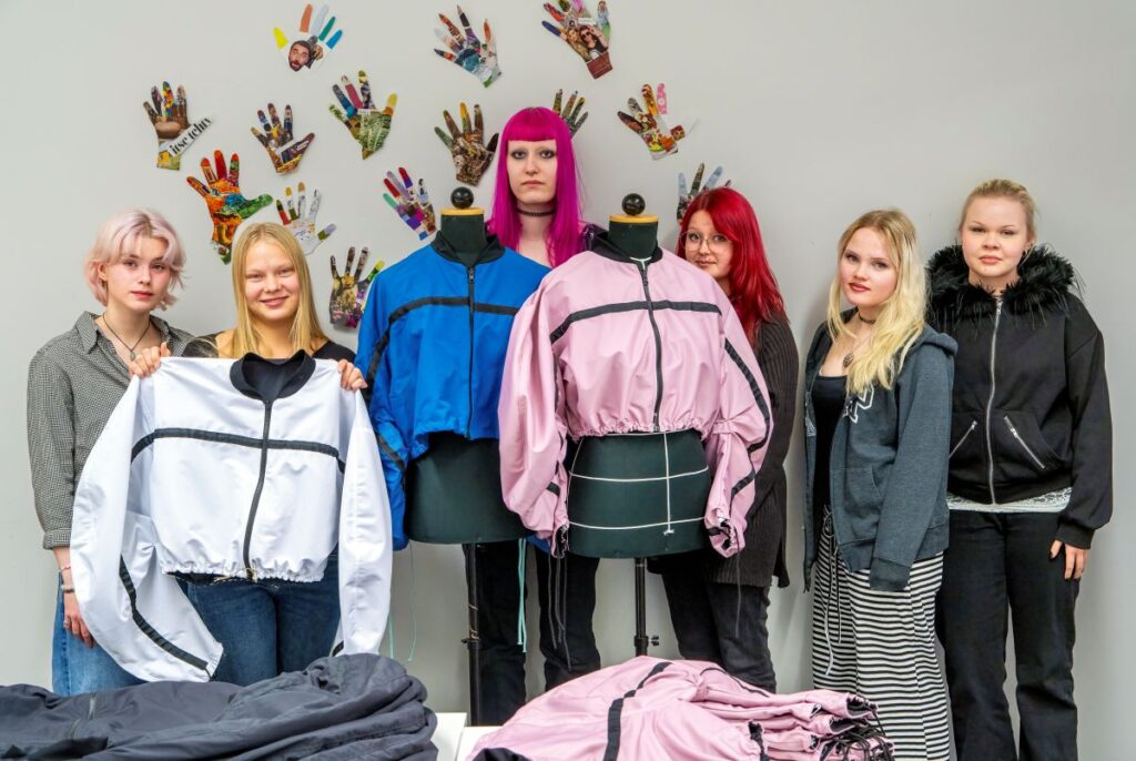 Ryhmä nuoria seisoo vaatetelineiden edessä, joilla on takkeja. Taustalla on koristeellisia kädenjälkiä seinällä.