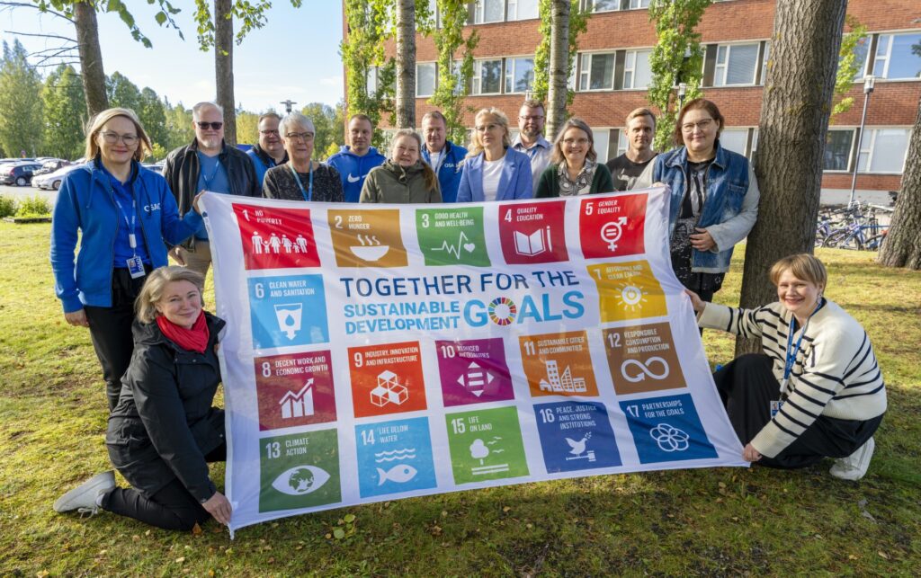 Ryhmä ihmisiä kokoontuneena ulkona pitäen Sustainable Development Goals -banderollia, jossa on 17 eri värikästä ruutua, ja jokaisessa ruudussa on erilainen tavoite. Taustalla on tiilirakennus ja puita.