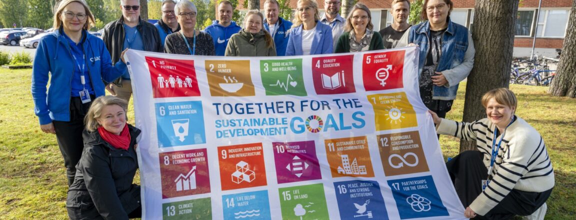 Ryhmä ihmisiä kokoontuneena ulkona pitäen Sustainable Development Goals -banderollia, jossa on 17 eri värikästä ruutua, ja jokaisessa ruudussa on erilainen tavoite. Taustalla on tiilirakennus ja puita.
