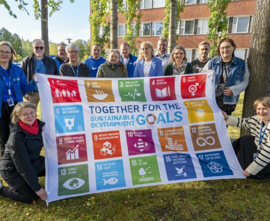 Ryhmä ihmisiä kokoontuneena ulkona pitäen Sustainable Development Goals -banderollia, jossa on 17 eri värikästä ruutua, ja jokaisessa ruudussa on erilainen tavoite. Taustalla on tiilirakennus ja puita.