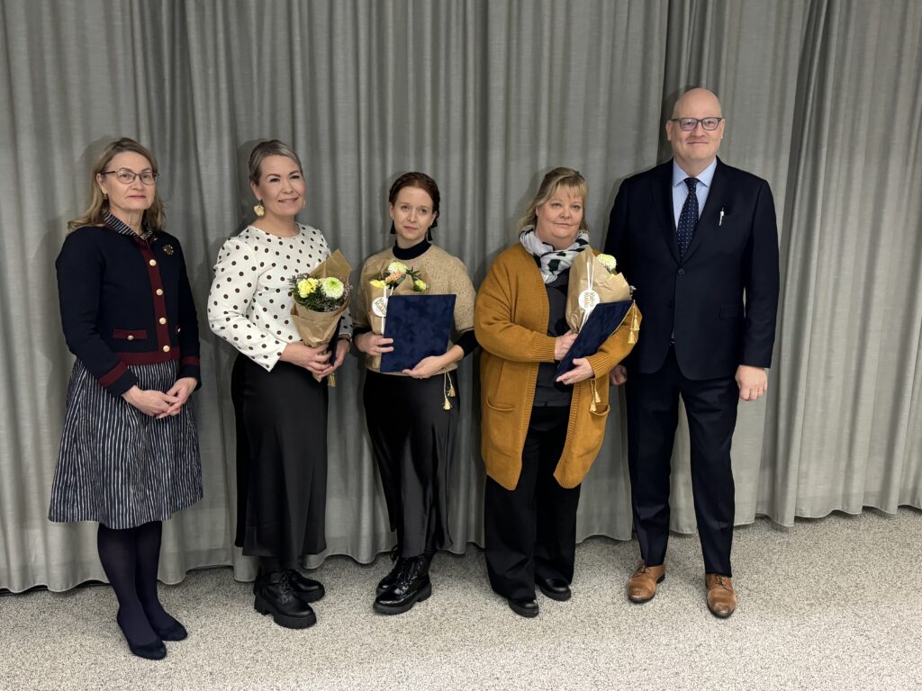 Viisi henkilöä seisoo rivissä harmaan verhon edessä. Kolmella heistä on kukkakimput käsissään. Henkilöt ovat pukeutuneet muodollisesti.