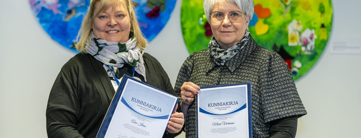 Kaksi naista pitelee kunniakirjoja värikkäiden pyöreiden taideteosten edessä.