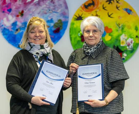 Kaksi naista pitelee kunniakirjoja värikkäiden pyöreiden taideteosten edessä.