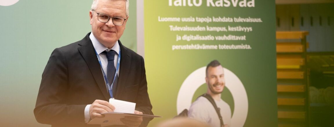 Mies seisoo OSAO:n mainosjulisteen edessä hymyillen. Julisteessa lukee 'Taito kasvaa', ja se korostaa tulevaisuuden kampusta, kestävyyttä ja digitalisaation merkitystä.