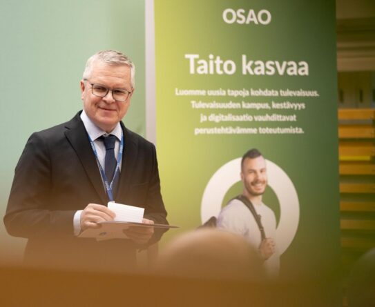 Mies seisoo OSAO:n mainosjulisteen edessä hymyillen. Julisteessa lukee 'Taito kasvaa', ja se korostaa tulevaisuuden kampusta, kestävyyttä ja digitalisaation merkitystä.
