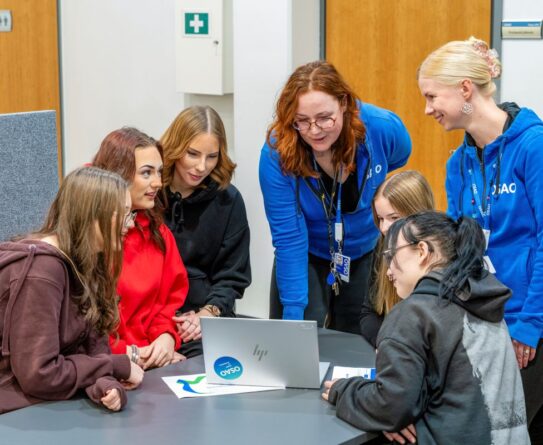 Ryhmä ihmisiä katsoo yhdessä kannettavaa tietokonetta. Kaksi henkilöä seisoo ja neljä muuta istuu pöydän ympärillä. Useimmat heistä pitävät yllään huppareita.