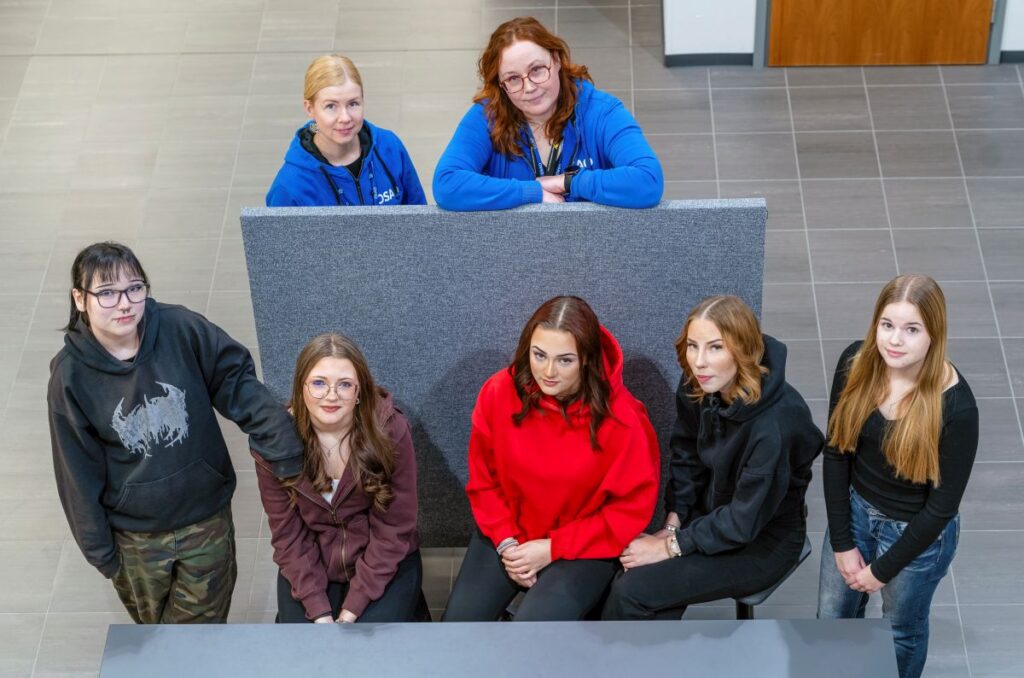 Seitsemän henkilöä istuu ja seisoo ryhmässä sisätiloissa. Kaksi henkilöä istuu harmaan sermin takana, ja viisi muuta on sermin edessä. He ovat pukeutuneet arkipäiväisiin vaatteisiin, kuten huppareihin ja farkkuihin. Taustalla näkyy harmaa lattia ja muutama ovi.