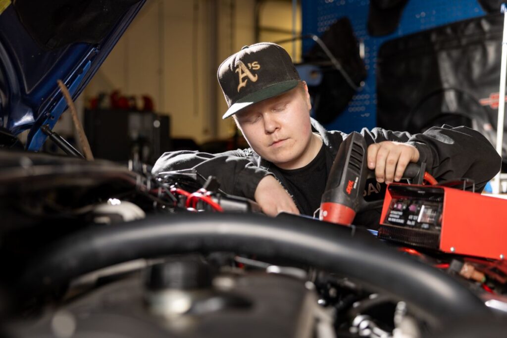 Henkilö työskentelee auton konepellin alla, käyttäen sähkötyökalua. Hänellä on päällään musta lippalakki, jossa on kirjain A.