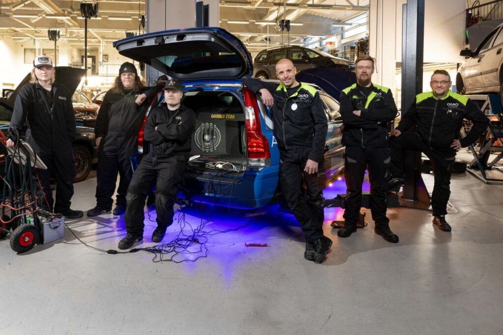 Kuusi henkilöä poseeraa auton ympärillä autotallissa. Auton takaluukku on auki, ja sisällä näkyy Ground Zero -äänijärjestelmä. Henkilöt ovat pukeutuneet tummiin työvaatteisiin, ja autosta loistaa sininen valo lattialle.