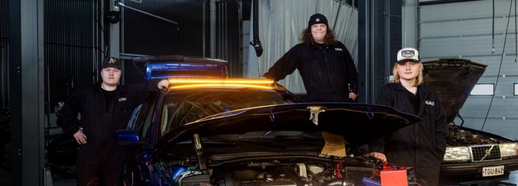 Kolme henkilöä seisoo autotallissa sinisen auton ympärillä, jonka konepelti on auki. Autossa on kirkkaat, oranssit valot etuosassa. Henkilöt ovat pukeutuneet tummiin vaatteisiin ja lippalakkeihin.