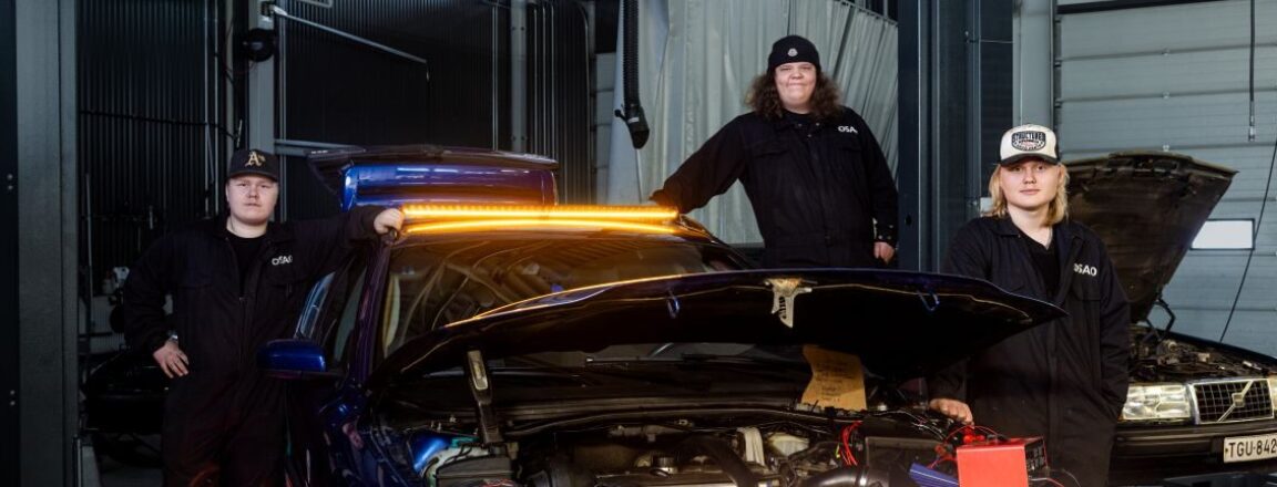 Kolme henkilöä seisoo autotallissa sinisen auton ympärillä, jonka konepelti on auki. Autossa on kirkkaat, oranssit valot etuosassa. Henkilöt ovat pukeutuneet tummiin vaatteisiin ja lippalakkeihin.