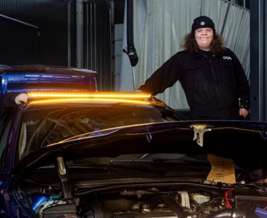 Kolme henkilöä seisoo autotallissa sinisen auton ympärillä, jonka konepelti on auki. Autossa on kirkkaat, oranssit valot etuosassa. Henkilöt ovat pukeutuneet tummiin vaatteisiin ja lippalakkeihin.