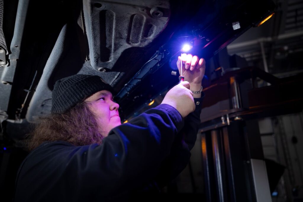 Henkilö korjaa ajoneuvon pohjaa autotallissa käyttäen työkalua ja valaisee työskentelyaluetta kirkkaalla valolla. Hänellä on yllään musta pipo ja tumma asu.