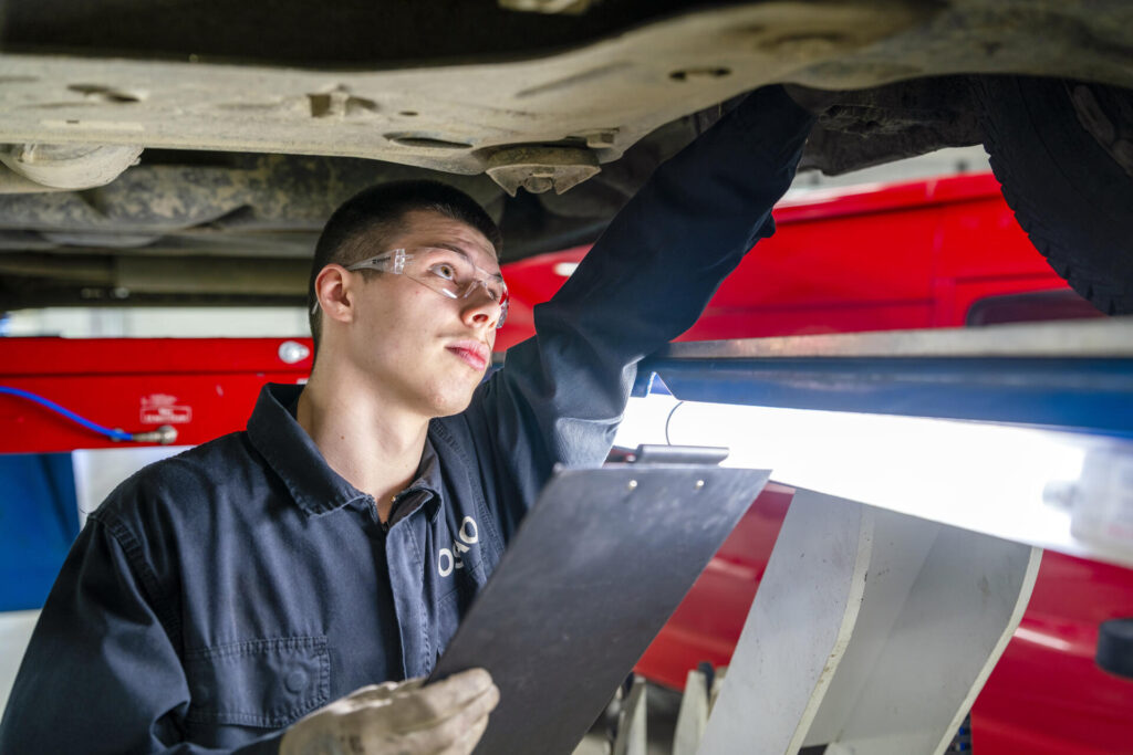Automekaanikko tarkastaa auton alustaa. Auto on ajoneuvonosturin päällä. Automekaanikolla on suojalasit ja käsissään kirjoitusalusta.