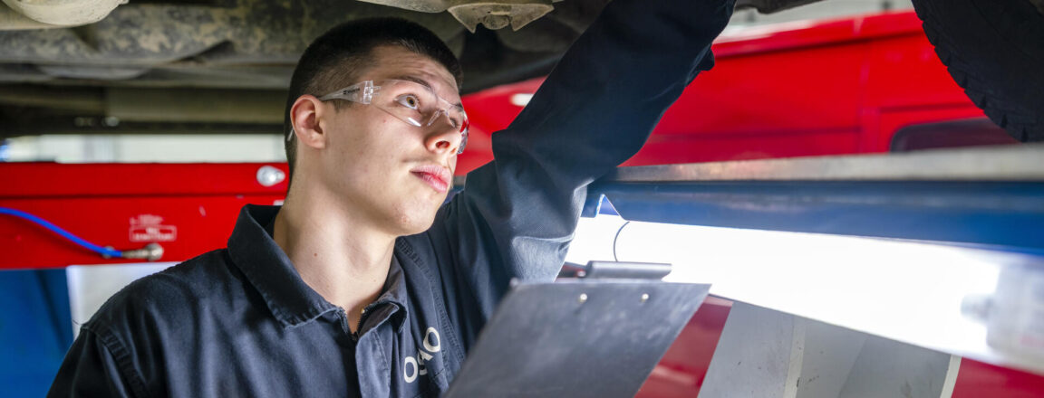 Automekaanikko tarkastaa auton alustaa. Auto on ajoneuvonosturin päällä. Automekaanikolla on suojalasit ja käsissään kirjoitusalusta.
