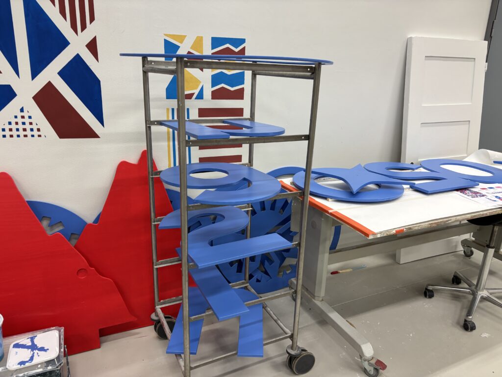 Työtila, jossa on metallinen hyllykkö täynnä sinisiä puulevyjä, jotka on muotoiltu numeroiksi. Taustalla seinällä on värikkäitä geometrisia kuvioita ja pöydällä lisää sinisiä muotoja.