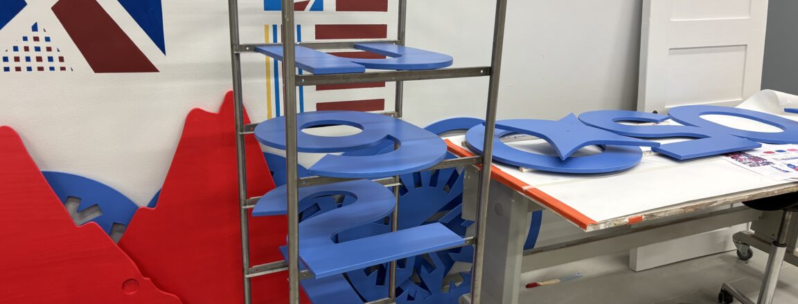 Työtila, jossa on metallinen hyllykkö täynnä sinisiä puulevyjä, jotka on muotoiltu numeroiksi. Taustalla seinällä on värikkäitä geometrisia kuvioita ja pöydällä lisää sinisiä muotoja.