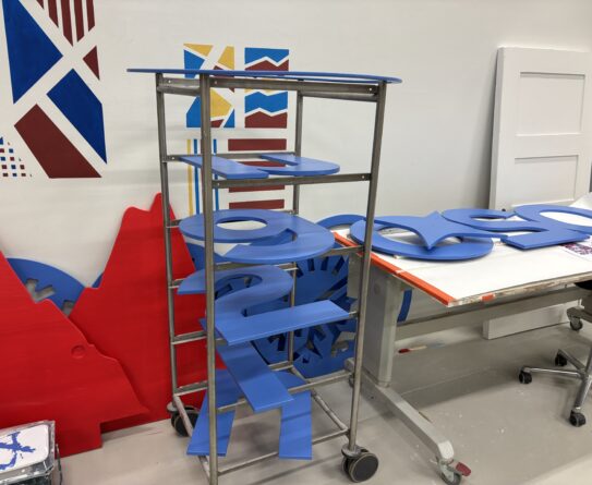 Työtila, jossa on metallinen hyllykkö täynnä sinisiä puulevyjä, jotka on muotoiltu numeroiksi. Taustalla seinällä on värikkäitä geometrisia kuvioita ja pöydällä lisää sinisiä muotoja.