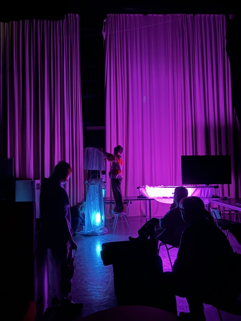 Hämärä studiotila, jossa on violetti valaistus. Keskellä on henkilö seisomassa tuolin päällä ja testaamassa tekstiilistä ja metallikehikosta koostuvaan rakennelmaan tulevaa valaistusta. Etualalla istuu ihmisiä seuraamassa tilannetta. Suuret verhot roikkuvat taustalla ja valaistuksen väri antaa tilaan mystisen tunnelman.